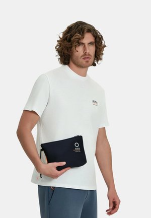 Uomo con capelli ricci indossa una camicia bianca a maniche corte e pantaloni blu, tenendo una pochette nera con cerniera e logo bianco all'altezza della vita.
