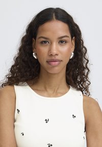 Donna con capelli ricci scuri che indossa orecchini a cerchio d'argento e una canottiera bianca con piccoli motivi floreali neri, rivolta in avanti.