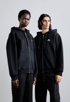 Čierne zip-up mikiny s prednými vreckami, s malým logom na hrudi. Textúrované čierne nohavice s obehovými vzormi.
