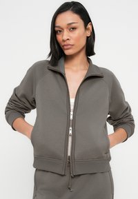 Ζακέτα με φερμουάρ σε απαλό ελαιδί πράσινο, κατασκευασμένη από μαλακό ύφασμα. Διαθέτει raglan μανίκια, ριμπ μανσέτες και ρυθμιζόμενο τελείωμα με κορδόνι για καλύτερη εφαρμογή.