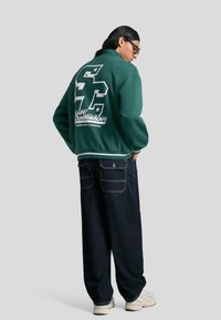 Jachetă verde cu fermoar și logo alb cu text pe spate. Purtați cu pantaloni de denim închis și pantofi deschiși. Material neted și croială lejeră.