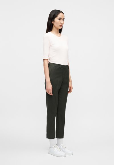 Νεαρή γυναίκα που στέκεται πλαγίως φορώντας ένα ανοιχτό ροζ κοντομάνικο τοπ, στενά σκούρα πράσινα παντελόνια, λευκές κάλτσες και λευκά αθλητικά παπούτσια.