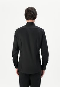 Cămașă neagră cu mâneci lungi, cu un design ajustat, guler mic, cusături pe spate pentru conturare și mansete cu nasturi. Textură netedă, detalii minime.