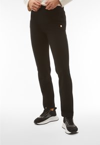 Black knit pants with a textured design and side pockets, featuring a small gold logo accent. Paired with black patterned sneakers.