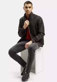 Czarna lekka kurtka z zamkami i stójką, w zestawieniu z czerwoną koszulą i ciemnymi dżinsami. Model siedzący na białym sześcianie.