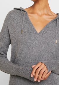 Lichtgrijze sweater met een V-hals, capuchon, geribde manchetten en trekkoorddetails van goudkleurig beslag. Zacht, geweven breisel.