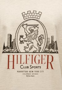 Μπεζ βαμβακερό t-shirt με μαύρο γραφικό λιονταριού που κρατά σπαθί, στέμμα ως διακριτικό και ορίζοντα πόλης, με κόκκινο κείμενο "HILFIGER CLUB SPORTS".