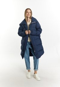 Geacă puffer albastră închisă cu guler înalt, închidere cu fermoar și manșete elastice. Purtați deasupra unui tricou ușor, asortată cu blugi răsuciți și teniși albi.