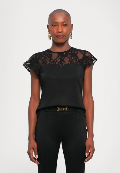 Woman with shaved head wearing black lace top and black pants with gold belt detail, gold hoop earrings, standing against white background.