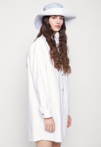 Vestido camiseiro branco oversized com fechamento de botões e mangas longas. Combinado com um chapéu branco de aba larga com uma faixa azul às riscas.