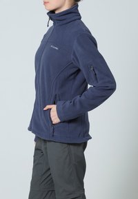 Tummansininen fleece-takki, jossa on korkea kaulus, vetoketjulliset sivutaskut ja pieni käsitasku. Takki on sileätekstuurinen ja istuva muotoilu.