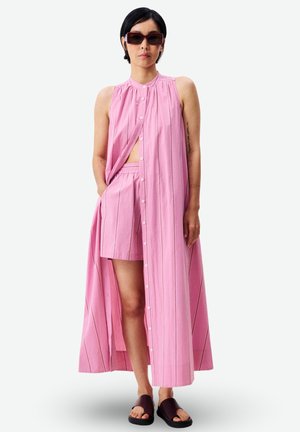 Femme portant une robe longue rose sans manches boutonnée sur des shorts assortis, des lunettes de soleil foncées et des sandales noires à enfiler, debout devant un fond blanc.