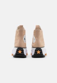 Para beżowych adidasów wysokich z białymi środkami, czarnymi podeszwami z pomarańczową gwiazdą i okrągłymi naszywkami z logo po bokach.