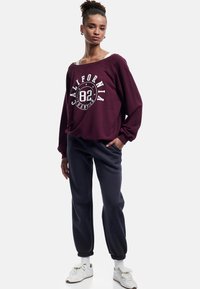 Sudadera oversize color marrón oscuro con estampado gráfico en blanco, combinada con joggers azul marino y zapatillas blancas con tiras de velcro.