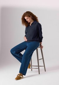 Pulover lung cu mâneci, de culoare albastră închisă, cu guler, asociat cu pantaloni din denim cu croi larg. Adidași maro cu accente albe completează ținuta casual.