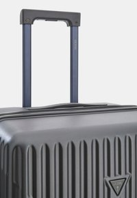 Grijze harde koffer met verticale ribbeltextuur, ritssluiting en een uitschuifbare donkerblauwe handgreep met het merkenlogo.