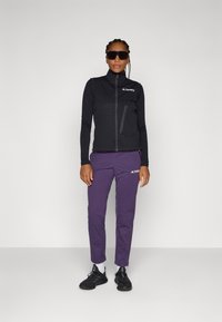 Black zip-up vest with textured design, paired with purple pants. Both items feature the Terrex logo. Black sneakers complete the outfit.