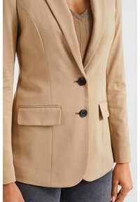 Blazer entallado color marrón claro con solapas de muesca, dos botones negros y bolsillos laterales, confeccionado con una tela suave de textura sutil.