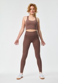 Barna sportfelső és magas derekú leggings, rugalmas anyagból, sima textúrával és minimális varrási részletekkel. Fehér sportcipők egészítik ki a megjelenést.