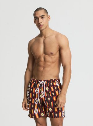Uomo senza maglietta che indossa pantaloncini da bagno colorati con motivi geometrici e coulisse, in piedi davanti a uno sfondo chiaro e uniforme.