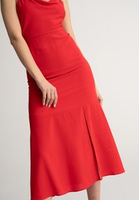 Czerwona, bezrękawna sukienka z gładkiego materiału, z dopasowaną górą i zwiewną spódnicą z rozcięciem z boku. Prosty, elegancki design.