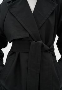 Manteau noir en trench avec un grand col, des manches larges, une ceinture à la taille et des poches. Confectionné en tissu lisse avec de subtils détails de couture.