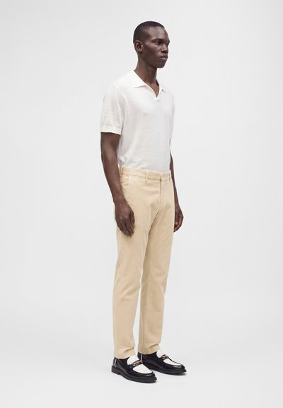 Homme debout portant un polo blanc à manches courtes, un pantalon beige, des chaussettes blanches et des mocassins noirs et blancs sur un fond uni.