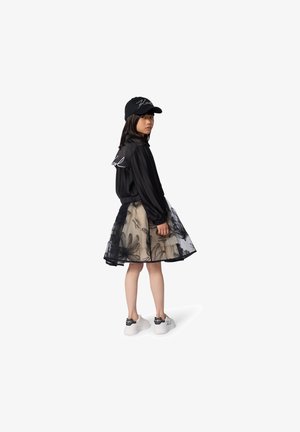 Veste bomber rayée noire avec logo en script, jupe en tulle à volants avec motif floral, et baskets blanches avec détails de nœuds noirs.
