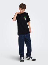 Pojke klädd i en svart t-shirt med tryck, lösa mörkblå jeans och svartvita sneakers, poserar med ena handen nära halsen.