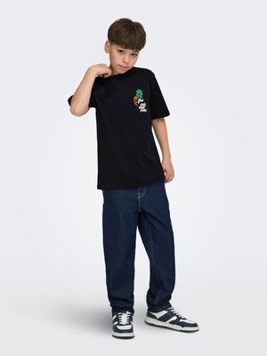 Jongen met een zwart T-shirt met een grafisch ontwerp, losse donkerblauwe spijkerbroek en zwart-witte sneakers, poserend met één hand bij zijn nek.