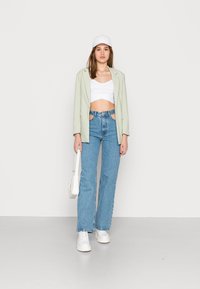 Blazer verde claro, top cropped branco, jeans azuis de cintura alta com recortes, sapatilhas brancas e um boné branco; acessórios minimalistas.