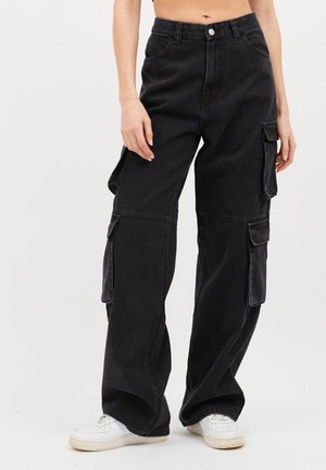 Persona que lleva pantalones cargo negros de tiro alto con múltiples bolsillos y zapatillas blancas, de pie contra un fondo liso.