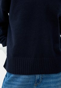Pull en laine bleu marine avec un ourlet et des poignets côtelés, à la texture lisse. Associé à un jean en denim bleu clair.