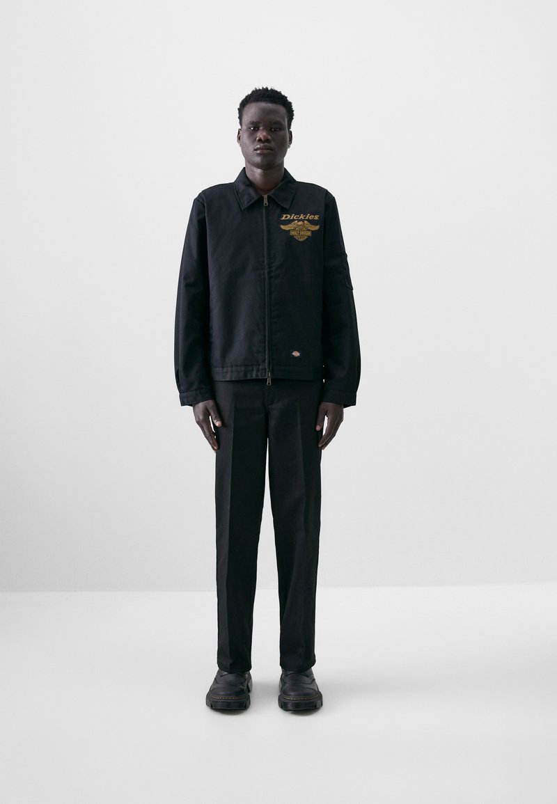 Black jacket with gold "Dickies" logo, zip closure, and side pockets, paired with matching black pants and chunky black shoes.