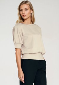 Beżowa bluzka z krótkim rękawem o luźnym kroju, z bufkami i dopasowanym dołem. Gładka tekstura z okrągłym dekoltem.