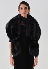 Wrap in pelliccia sintetica nera con una texture soffice e maniche larghe, dettagliato con un'accortezza in raso nero a forma di fiocco sul collo.