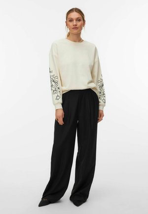 Donna che indossa un top crema a maniche lunghe con motivi floreali neri sulle maniche e pantaloni neri larghi e ampi, in piedi su uno sfondo bianco semplice.