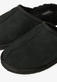 Mocasines de ante negro con un suave forro interior, que presentan un detalle de costura y una suela de goma negra texturizada. La marca es visible en la suela.