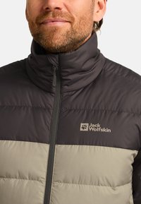 Veste matelassée en gris foncé et beige clair, avec un col montant et une fermeture éclair intégrale, arborant le logo Jack Wolfskin sur la poitrine.