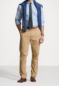 Camicia azzurro chiaro con maniche arrotolate, cravatta a righe verde e blu navy, cintura in pelle marrone, pantaloni khaki e maglione a trecce blu navy a drappo.