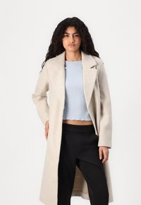Lang beige ullkjole med spiss krage, rett snitt og frontlommer, båret over en lys blå teksturert topp og svarte bukser.