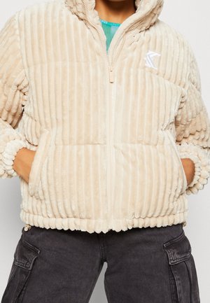 Henkilö pukeutuneena beigeen ribbineulottuun pehmeään vetoketjutakkiin, kädet taskuissa ja yllään tummat cargo-housut, näkyvissä kaulasta lantiolle.