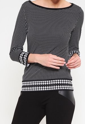Femme portant un haut à manches longues noir et blanc à petits carreaux avec poignets et ourlet à motif pied-de-poule, associé à un pantalon noir.