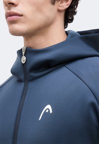 Κοντινό πλάνο ενός ατόμου που φοράει μια καφέ-μπλε φούτερ με φερμουάρ και ένα λευκό στυλιζαρισμένο λογότυπο στο στήθος και στρογγυλό τράβηγμα φερμουάρ.