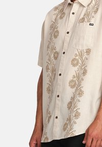 Camisa de manga corta beige con bordado floral en dorado, que cuenta con un bolsillo en el pecho, cierre de botones en la parte frontal y un corte relajado.