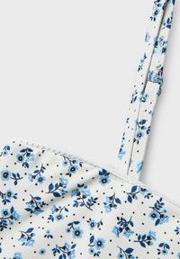 Blommig crop top med vit bakgrund, blå blommor och svarta prickar, justerbara axelband och spetskant längs kanten.