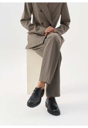 Personne portant un costume marron, assise les jambes croisées, les mains jointes, et des chaussures en cuir noir à lacets visibles sur un fond blanc.