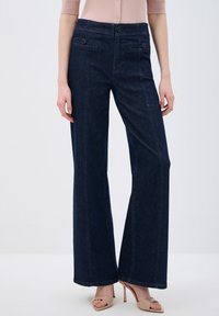 Jeans de mezclilla de pierna ancha en azul oscuro con bolsillos frontales, detalles de costura sutiles y una textura suave; combinados con sandalias de tacón alto beige.