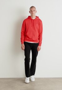 Felpa rossa con cappuccio, tasca frontale, cordino e patch con logo, abbinata a pantaloni neri e sneakers bianche. Sullo sfondo bianco.