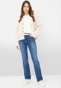 Beige vest met knopen, wit T-shirt, blauwe uitgerafelde jeans en witte sneakers. Gladde textuur en casual pasvorm worden overal benadrukt.